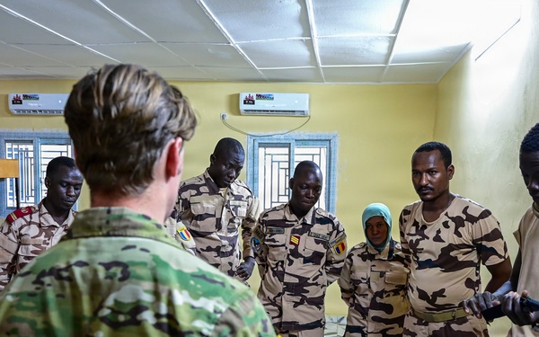 A military training or briefing session is taking place indoors, where a soldier in green multicam uniform addresses a group of personnel wearing desert camouflage uniforms bearing what appears to be the Chadian flag patch. The setting appears to be a classroom or meeting room with yellow walls and air conditioning units. The group is attentively listening to the briefer who is gesturing while speaking.