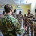 A military training or briefing session is taking place indoors, where a soldier in green multicam uniform addresses a group of personnel wearing desert camouflage uniforms bearing what appears to be the Chadian flag patch. The setting appears to be a classroom or meeting room with yellow walls and air conditioning units. The group is attentively listening to the briefer who is gesturing while speaking.