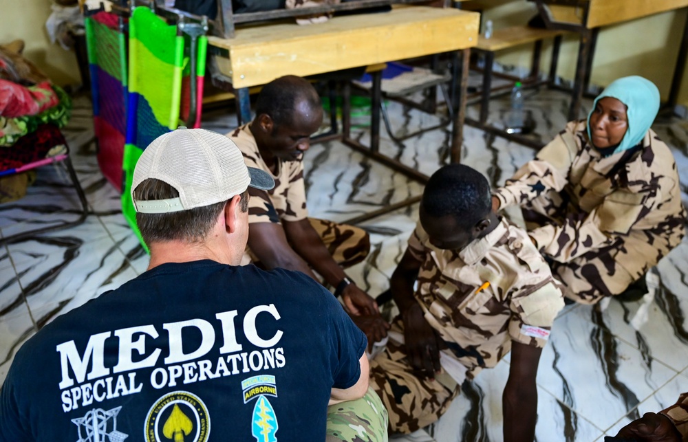A Special Operations Medic wearing a black 'MEDIC SPECIAL OPERATIONS' t-shirt is conducting what appears to be a medical training session with local military personnel wearing desert camouflage uniforms. The group is seated on the floor of what looks like a classroom or training facility. Three local soldiers, including one woman wearing a teal hijab, are participating in the session.