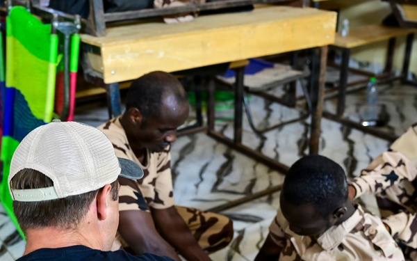 A Special Operations Medic wearing a black 'MEDIC SPECIAL OPERATIONS' t-shirt is conducting what appears to be a medical training session with local military personnel wearing desert camouflage uniforms. The group is seated on the floor of what looks like a classroom or training facility. Three local soldiers, including one woman wearing a teal hijab, are participating in the session.