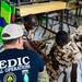 A Special Operations Medic wearing a black 'MEDIC SPECIAL OPERATIONS' t-shirt is conducting what appears to be a medical training session with local military personnel wearing desert camouflage uniforms. The group is seated on the floor of what looks like a classroom or training facility. Three local soldiers, including one woman wearing a teal hijab, are participating in the session.