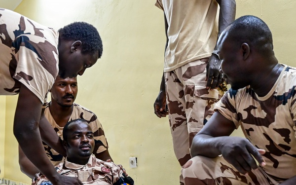 Several soldiers in matching desert camouflage uniforms are gathered around a seated colleague on an indoor floor. The scene appears to depict a first aid or combat casualty care training exercise, with one soldier simulating an injury indicated by white bandaging on his leg. The setting is an interior room with yellow walls and tiled flooring.