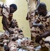 Several soldiers in matching desert camouflage uniforms are gathered around a seated colleague on an indoor floor. The scene appears to depict a first aid or combat casualty care training exercise, with one soldier simulating an injury indicated by white bandaging on his leg. The setting is an interior room with yellow walls and tiled flooring.