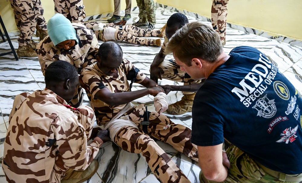 A Special Operations medic in a navy blue t-shirt is conducting a medical training exercise with military personnel wearing desert camouflage uniforms. The training appears to focus on casualty care, specifically bandaging and tourniquet application on a simulated wounded soldier's leg. The setting appears to be an indoor facility with a tiled floor.