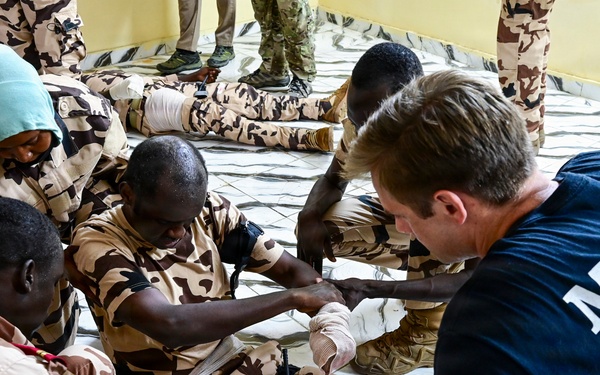 A Special Operations medic in a navy blue t-shirt is conducting a medical training exercise with military personnel wearing desert camouflage uniforms. The training appears to focus on casualty care, specifically bandaging and tourniquet application on a simulated wounded soldier's leg. The setting appears to be an indoor facility with a tiled floor.