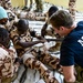 A Special Operations medic in a navy blue t-shirt is conducting a medical training exercise with military personnel wearing desert camouflage uniforms. The training appears to focus on casualty care, specifically bandaging and tourniquet application on a simulated wounded soldier's leg. The setting appears to be an indoor facility with a tiled floor.