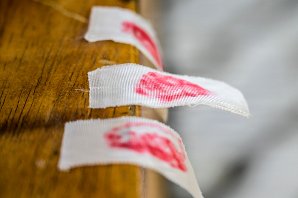 The image shows several small white fabric or woven labels attached along the edge of a wooden surface, each bearing a red printed mark or stamp. The labels appear to be sewn or affixed in a row along what looks like a wooden beam or furniture edge. The depth of field is shallow, with the middle label in sharpest focus.