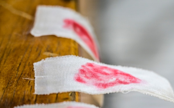 The image shows several small white fabric or woven labels attached along the edge of a wooden surface, each bearing a red printed mark or stamp. The labels appear to be sewn or affixed in a row along what looks like a wooden beam or furniture edge. The depth of field is shallow, with the middle label in sharpest focus.