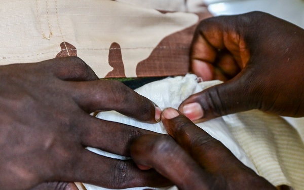 A close-up image showing two pairs of hands, with one person appearing to apply a bandage or dressing to another person's hand or finger. The subject wearing desert camouflage (tan/brown pattern) military uniform is receiving what appears to be first aid or medical treatment. A gold ring is visible on one of the hands administering care.