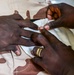 A close-up image showing two pairs of hands, with one person appearing to apply a bandage or dressing to another person's hand or finger. The subject wearing desert camouflage (tan/brown pattern) military uniform is receiving what appears to be first aid or medical treatment. A gold ring is visible on one of the hands administering care.