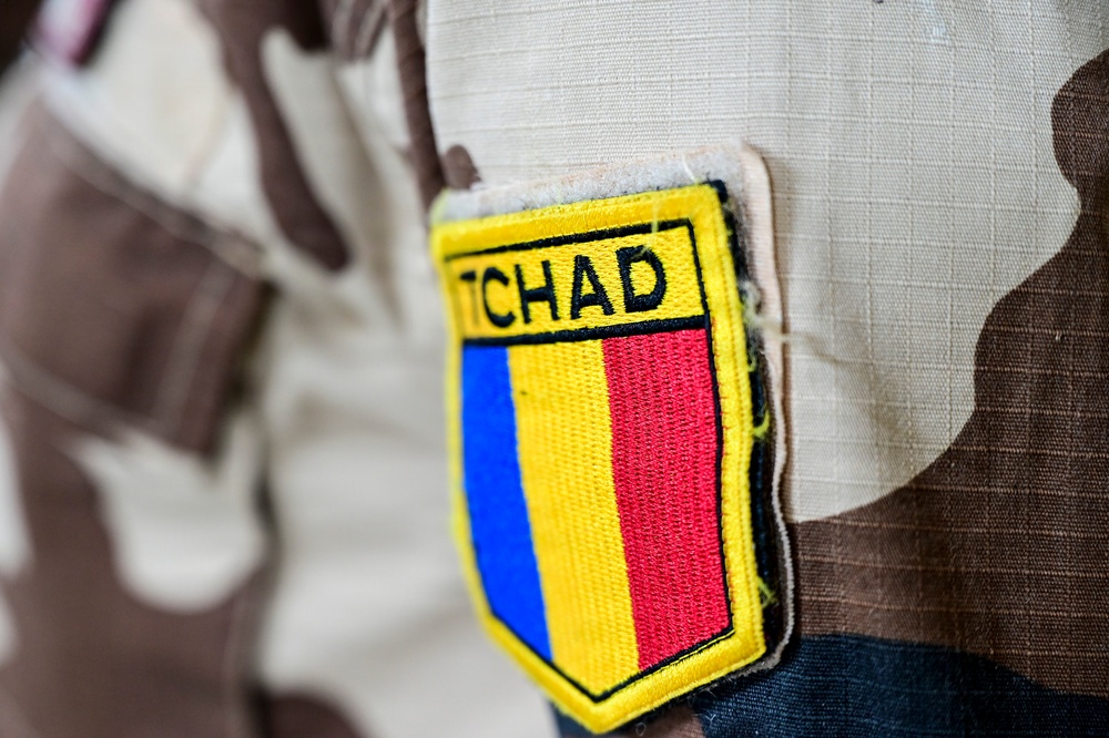 A close-up photograph of a military uniform sleeve patch reading 'TCHAD' (the French name for Chad). The patch displays the Chadian national flag colors — blue, yellow, and red — in a shield-shaped emblem with a yellow border. The uniform itself features a desert camouflage pattern typical of Sahelian military forces.