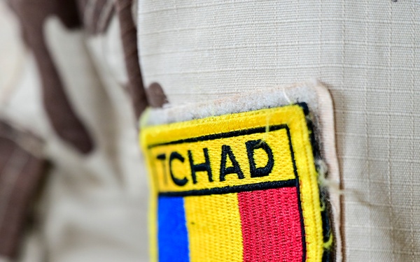 A close-up photograph of a military uniform sleeve patch reading 'TCHAD' (the French name for Chad). The patch displays the Chadian national flag colors — blue, yellow, and red — in a shield-shaped emblem with a yellow border. The uniform itself features a desert camouflage pattern typical of Sahelian military forces.