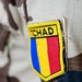 A close-up photograph of a military uniform sleeve patch reading 'TCHAD' (the French name for Chad). The patch displays the Chadian national flag colors — blue, yellow, and red — in a shield-shaped emblem with a yellow border. The uniform itself features a desert camouflage pattern typical of Sahelian military forces.