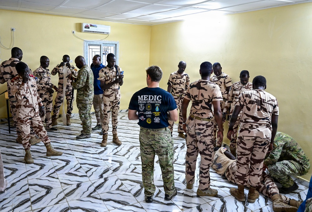 A Special Operations medic in a navy blue 'MEDIC SPECIAL OPERATIONS' t-shirt and multicam trousers is conducting a medical training session with a group of soldiers wearing desert camouflage uniforms. The training appears to take place inside a yellow-walled room with a marble-patterned tile floor, likely in an African country based on contextual indicators. A practical demonstration or scenario exercise is visible in the foreground-right where a soldier is kneeling over a simulated casualty.