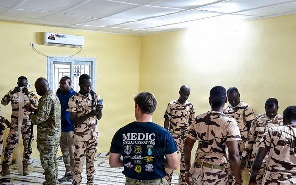 A Special Operations medic in a navy blue 'MEDIC SPECIAL OPERATIONS' t-shirt and multicam trousers is conducting a medical training session with a group of soldiers wearing desert camouflage uniforms. The training appears to take place inside a yellow-walled room with a marble-patterned tile floor, likely in an African country based on contextual indicators. A practical demonstration or scenario exercise is visible in the foreground-right where a soldier is kneeling over a simulated casualty.