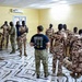 A Special Operations medic in a navy blue 'MEDIC SPECIAL OPERATIONS' t-shirt and multicam trousers is conducting a medical training session with a group of soldiers wearing desert camouflage uniforms. The training appears to take place inside a yellow-walled room with a marble-patterned tile floor, likely in an African country based on contextual indicators. A practical demonstration or scenario exercise is visible in the foreground-right where a soldier is kneeling over a simulated casualty.