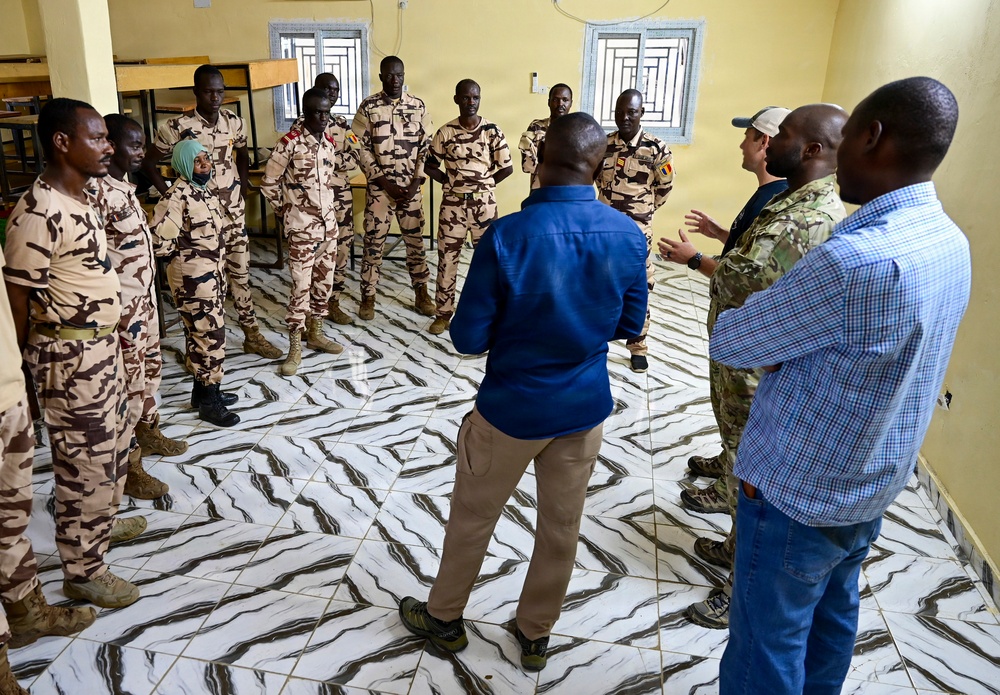 A group of uniformed military personnel and civilians are gathered in a circle inside a room with a distinctive marble-patterned tile floor and yellow walls. The military personnel are wearing desert camouflage uniforms with what appear to be Chadian flag patches, while several civilians in casual attire, including one individual in multicam/military-style clothing, appear to be engaged in a briefing or discussion. The setting suggests a training, coordination, or advisory meeting between local military forces and international personnel.