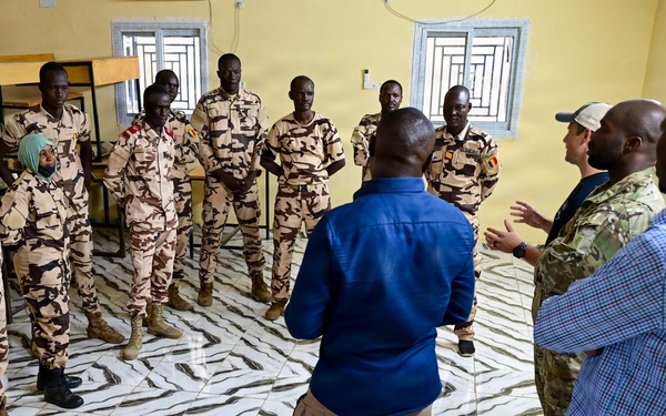 A group of uniformed military personnel and civilians are gathered in a circle inside a room with a distinctive marble-patterned tile floor and yellow walls. The military personnel are wearing desert camouflage uniforms with what appear to be Chadian flag patches, while several civilians in casual attire, including one individual in multicam/military-style clothing, appear to be engaged in a briefing or discussion. The setting suggests a training, coordination, or advisory meeting between local military forces and international personnel.
