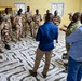 A group of uniformed military personnel and civilians are gathered in a circle inside a room with a distinctive marble-patterned tile floor and yellow walls. The military personnel are wearing desert camouflage uniforms with what appear to be Chadian flag patches, while several civilians in casual attire, including one individual in multicam/military-style clothing, appear to be engaged in a briefing or discussion. The setting suggests a training, coordination, or advisory meeting between local military forces and international personnel.