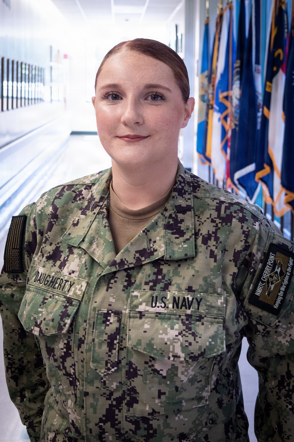 A U.S. Navy service member named Daugherty is photographed in a military corridor wearing the Navy Working Uniform (NWU) digital camouflage pattern. She is assigned to NMRTC Cherry Point, as indicated by the unit patch on her right sleeve. The setting appears to be an official military facility with a row of flags visible in the background.