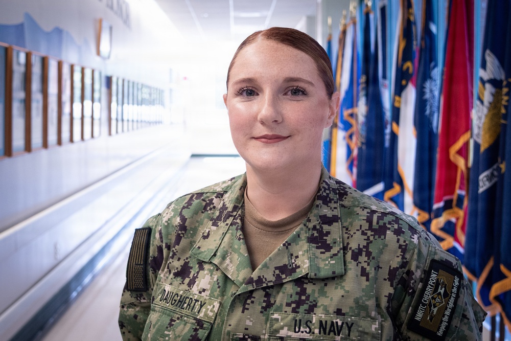 A U.S. Navy service member named Daugherty is photographed in a military facility hallway. She is wearing the Navy Working Uniform (NWU) with visible name tape reading 'DAUGHERTY' and 'U.S. NAVY' branch tape. The background features a row of military and state flags along with plaques or awards mounted on the wall.