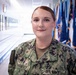 A U.S. Navy service member named Daugherty is photographed in a military facility hallway. She is wearing the Navy Working Uniform (NWU) with visible name tape reading 'DAUGHERTY' and 'U.S. NAVY' branch tape. The background features a row of military and state flags along with plaques or awards mounted on the wall.