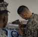 Two military personnel in MARPAT (Marine pattern) camouflage uniforms are engaged in what appears to be a supply inventory or documentation activity. One service member is actively writing on a clipboard or document while the other observes. Cases of bottled water and other supplies are visible in the background.