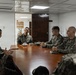 A group of military personnel in camouflage uniforms are seated around a wooden conference table in what appears to be a briefing or meeting room. The individuals appear to represent at least two different military organizations, identifiable by slightly different camouflage patterns. The setting suggests a formal military coordination or liaison meeting.