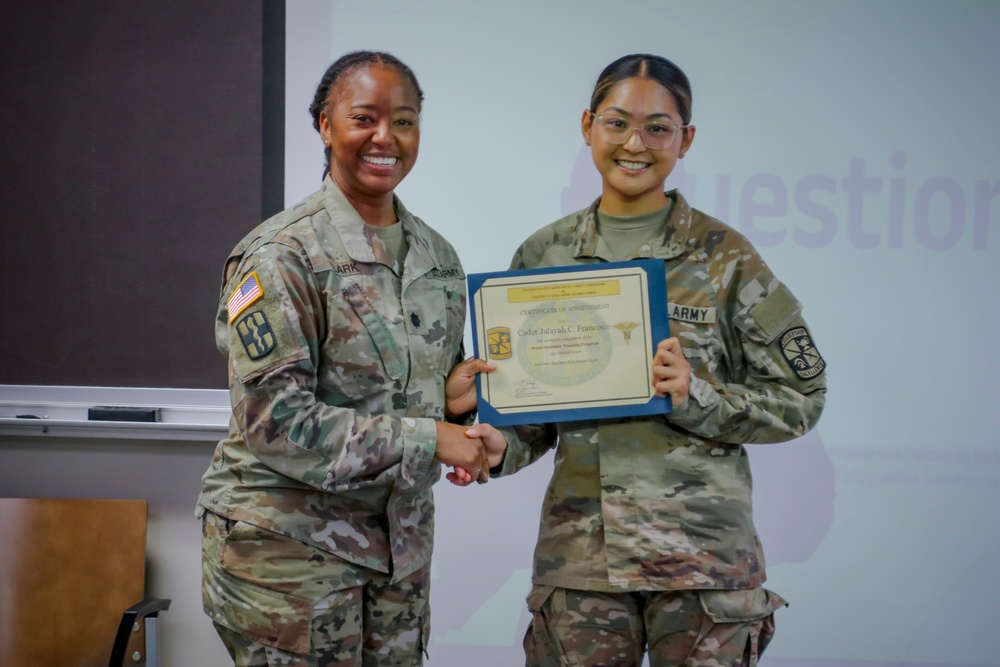 Two U.S. Army soldiers in Army Combat Uniforms (ACU/OCP pattern) pose together while holding a certificate. The soldier on the right appears to be receiving a Certificate of Achievement, with both individuals shaking hands. The ceremony appears to be taking place in a classroom or briefing room setting.