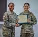 Two U.S. Army soldiers in Army Combat Uniforms (ACU/OCP pattern) pose together while holding a certificate. The soldier on the right appears to be receiving a Certificate of Achievement, with both individuals shaking hands. The ceremony appears to be taking place in a classroom or briefing room setting.