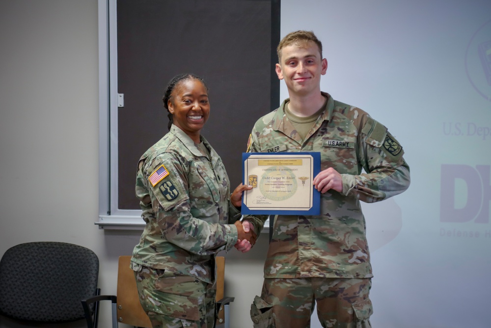 Two U.S. Army personnel in OCP (Operational Camouflage Pattern) uniforms are posing together during a certificate presentation ceremony. A female soldier is presenting a Certificate of Achievement to a male soldier identified by his name tape as 'EMLER'. Both are smiling and shaking hands while jointly holding the framed certificate.