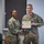 Two U.S. Army personnel in OCP (Operational Camouflage Pattern) uniforms are posing together during a certificate presentation ceremony. A female soldier is presenting a Certificate of Achievement to a male soldier identified by his name tape as 'EMLER'. Both are smiling and shaking hands while jointly holding the framed certificate.