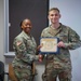 Two U.S. Army personnel in OCP (Operational Camouflage Pattern) uniforms are posing together during a certificate presentation ceremony. A female soldier is presenting a Certificate of Achievement to a male soldier identified by his name tape as 'EMLER'. Both are smiling and shaking hands while jointly holding the framed certificate.