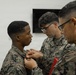 Three U.S. Marines in digital camouflage uniforms (MARPAT) are engaged in what appears to be a military decoration or insignia pinning ceremony. Two senior Marines are attaching something to the chest area of a younger Marine, suggesting a promotion or award ceremony. The setting appears to be an indoor military facility.