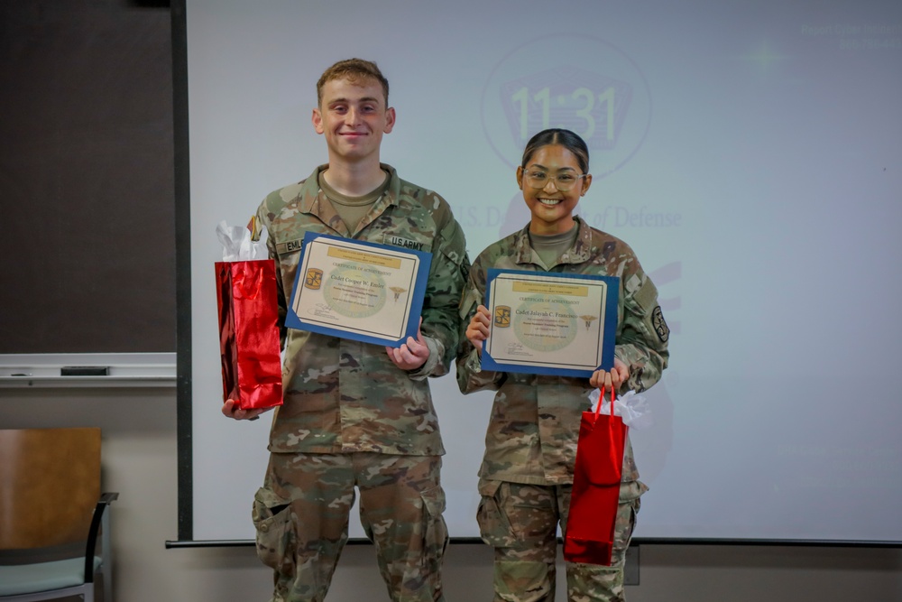 Two U.S. Army cadets in military camouflage uniforms pose together holding certificates of achievement and red gift bags. They are standing in front of a projection screen displaying what appears to be a U.S. Department of Defense logo. Both individuals are smiling, suggesting a celebratory or award ceremony occasion.