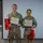 Two U.S. Army cadets in military camouflage uniforms pose together holding certificates of achievement and red gift bags. They are standing in front of a projection screen displaying what appears to be a U.S. Department of Defense logo. Both individuals are smiling, suggesting a celebratory or award ceremony occasion.