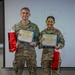 Two U.S. Army cadets in military camouflage uniforms pose together holding certificates of achievement and red gift bags. They are standing in front of a projection screen displaying what appears to be a U.S. Department of Defense logo. Both individuals are smiling, suggesting a celebratory or award ceremony occasion.