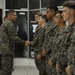 A senior military officer in digital camouflage uniform is shaking hands with junior personnel lined up in formation inside a building. The scene appears to depict a formal greeting or recognition ceremony, with multiple service members standing at attention in a row. All individuals are wearing matching digital camouflage uniforms consistent with U.S. Marine Corps MARPAT pattern.