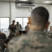 A military briefing or discussion session is taking place in what appears to be a room aboard a naval vessel or coastal facility, with large windows overlooking open water. A senior military figure, seen from behind in the foreground, is addressing a group of uniformed personnel seated in a semicircle. One service member is standing and appears to be speaking or asking a question.
