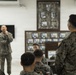 A military briefing or informal gathering is taking place indoors, with a smiling speaker in camouflage uniform addressing a group of seated and standing military personnel. The setting appears to be a common room or meeting hall on a military installation, decorated with framed photographs on the wall. The atmosphere appears relaxed and conversational rather than a formal parade or operational setting.