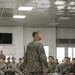 A military briefing or address is taking place indoors, with one senior service member standing and speaking to a large group of seated personnel. All individuals are wearing MARPAT (Marine Pattern) digital camouflage uniforms consistent with the United States Marine Corps. The setting appears to be a multipurpose room or meeting hall on a military installation.