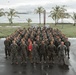 A large group of military personnel in camouflage uniforms poses for a formal unit photograph on a wet tarmac or parade ground. A red guidon flag is visible at the center front of the formation, consistent with U.S. Marine Corps unit markings. The backdrop features palm trees, a grassy coastal area, and a body of water with what appears to be a port or harbor in the distance.