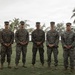Five military personnel in matching camouflage uniforms stand in a formal group pose on a grass field. They appear to be U.S. Marines based on their MARPAT (Marine Pattern) digital camouflage uniforms and cover caps. The setting appears to be a tropical coastal location, likely a military installation, with palm trees and water visible in the background.