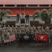 A large group of U.S. Marines in woodland MARPAT camouflage uniforms poses for a formal unit photograph in front of a military monument featuring bronze soldier statues. The group displays three flags: the American flag, a black unit guidon labeled 'BLACKFOOT' with a skull insignia, and the United States Marine Corps flag. The setting appears to be an overseas military installation, likely in a tropical region based on the palm trees and red-roofed building.