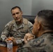 Two military personnel are seated across from each other at a table in what appears to be a conference or briefing room. The individual facing the camera wears a camouflage uniform with a name tape reading 'GONZALEZ' and visible rank insignia, while the second individual, seen from behind, wears a different camouflage pattern consistent with U.S. Marine Corps MARPAT. They appear to be engaged in a formal or semi-formal discussion.