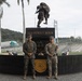 Two military personnel in different camouflage uniforms are posing in front of a military monument inscribed with 'DIOS PATRIA Y HONOR' (God, Homeland and Honor in Spanish). The monument features a bronze soldier statue on top of a dark stone pedestal, flanked by two historic cannons. The setting appears to be a Latin American military installation, likely in Ecuador or a similar Spanish-speaking country.