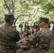Two U.S. Marines in MARPAT digital camouflage uniforms are engaged in conversation in a forested environment, facing away from the camera. A larger group of personnel is visible in the background, gathered near large tree roots in what appears to be a jungle or dense woodland setting. The scene suggests a field training exercise or operational briefing in a tropical environment.