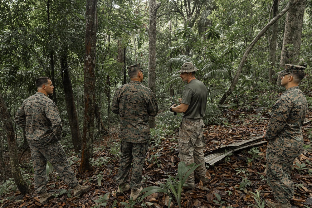 Four military personnel in a tropical jungle setting appear to be engaged in a briefing or discussion. Three individuals are wearing MARPAT (Marine pattern) camouflage uniforms, while one wears a plain olive/green t-shirt with camouflage trousers and a boonie hat. A piece of corrugated metal sheeting is visible on the ground among fallen leaves.