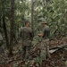 Four military personnel in a tropical jungle setting appear to be engaged in a briefing or discussion. Three individuals are wearing MARPAT (Marine pattern) camouflage uniforms, while one wears a plain olive/green t-shirt with camouflage trousers and a boonie hat. A piece of corrugated metal sheeting is visible on the ground among fallen leaves.