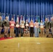 A formal awards ceremony taking place on a stage at Fort Campbell military installation, where several civilians are being recognized with certificates by military personnel. A group of approximately 11 individuals — including uniformed Army soldiers and civilian recipients — pose together on a wooden stage in front of a backdrop of military unit flags and Fort Campbell banners. Six civilians are visibly holding red-bordered certificates or award documents.